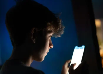 نمای جانبی گوشی هوشمند در محیط کم‌نور با صفحه‌نمایش روشن، نمایش محتوای اپلیکیشن در دست کاربر و نور صفحه به‌عنوان منبع نور اصلی صحنه