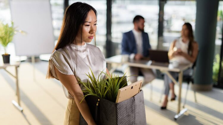 زنی با چهرهای غمگین در حال حمل جعبه وسایل شخصی خود پس از اخراج از محیط کار.