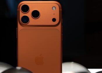 نمای نزدیک به پشت یک گوشی نارنجی رنگ (ظاهراً iPhone 17 Pro) با سه لنز دوربین بزرگ.