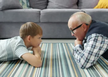 یک کودک و یک فرد مسن در حال دراز کشیدن روی فرش راه‌راه در اتاق نشیمن و گفت‌وگو در مقابل مبل خاکستری