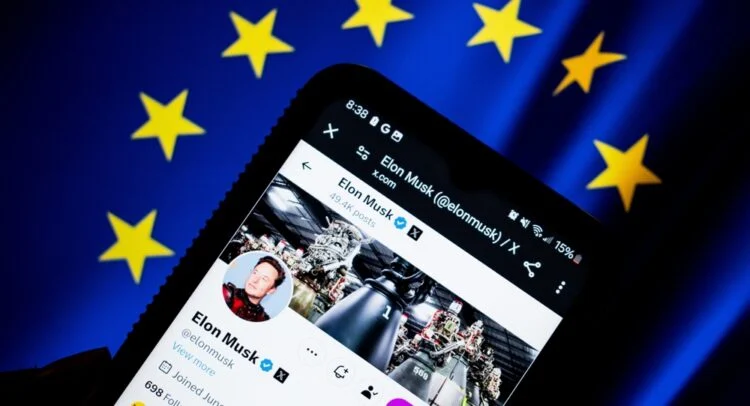 گوشی هوشمندی که نمایه کاربری ایلان ماسک در X را نشان می‌دهد، با پرچم اتحادیه اروپا و ستاره‌های آن در پس‌زمینه.