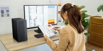 زنی در حال کار با کامپیوتر ایسوس ExpertCenter و بررسی نقشه‌های معماری روی میز کار.