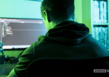 تصویر نمادین از یک هکر با هودی که در اتاقی با نور سبز جلوی کامپیوتر نشسته است.