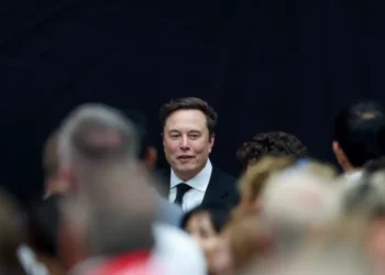 ایلان ماسک با کت و شلوار و چهره‌ای خندان در مرکز تصویر، در میان جمعیتی که فوکوس ندارند (احتمالا جلسه xAI).