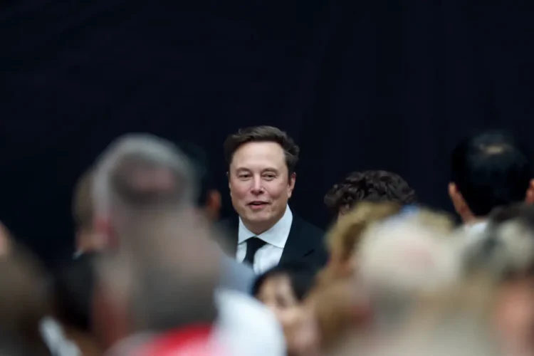 ایلان ماسک با کت و شلوار و چهرهای خندان در مرکز تصویر، در میان جمعیتی که فوکوس ندارند (احتمالا جلسه xAI).