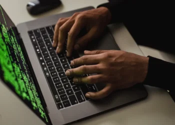 نمای نزدیک از لپ‌تاپی با نمایش کدهای سبزرنگ روی صفحه‌نمایش، قسمتی از یک محیط کاری با فضای اداری مدرن