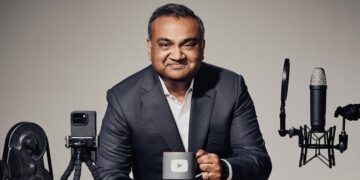 نیل موهان (مدیرعامل یوتیوب) نشسته کنار تجهیزات ضبط و ماگ با لوگوی یوتیوب.