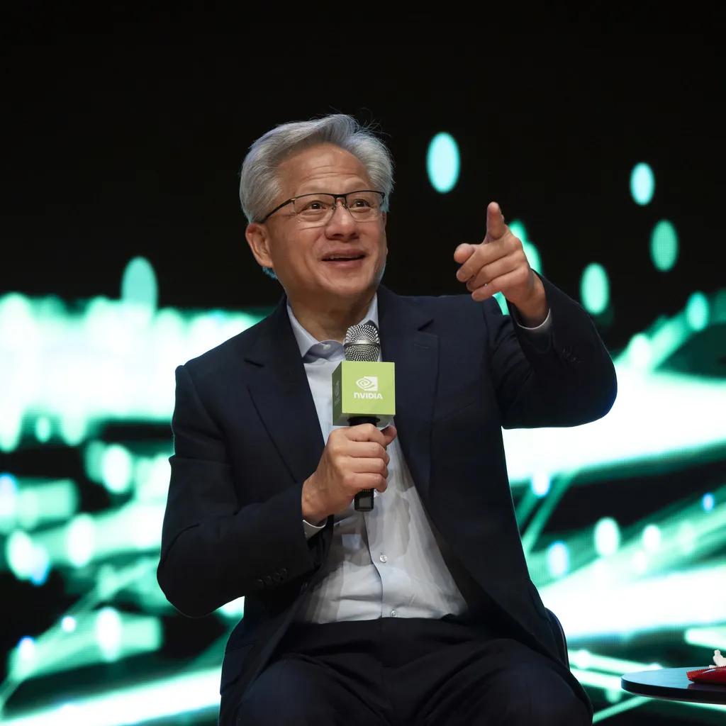 جنسن هوانگ، مدیرعامل انویدیا، با کت و شلوار سرمه‌ای در حال سخنرانی با میکروفون و اشاره به سمت جمعیت.
