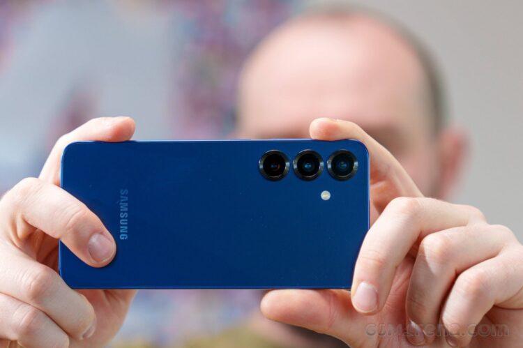 شخصی در حال عکاسی با یک گوشی سامسونگ گلکسی آبی رنگ که لنزهای سهگانه دوربین پشت آن مشخص است.