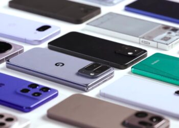 نمای نزدیک از ده‌ها گوشی هوشمند پرچمدار و میان‌رده سال ۲۰۲۵ که به صورت منظم روی میز سفید چیده شده‌اند.