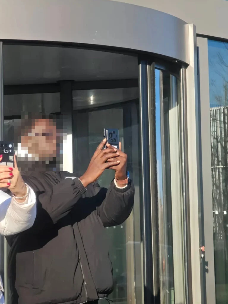 فردی که گوشی را در مقابل یک ساختمان شیشهای برای عکاسی بالا گرفته است؛ چهره کاربر شطرنجی شده و پشت گوشی دیده میشود.