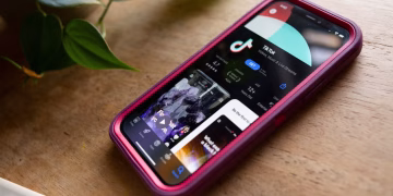 گوشی آیفون روی میز که صفحه دانلود تیک‌تاک در اپ‌استور را نشان می‌دهد.