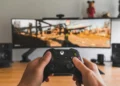 نمای اول شخص گیمینگ با کنترلر ایکس‌باکس روی میز، نمایشگر بزرگ با تصویر بازی اکشن، اسپیکرهای استودیویی و تزئینات شلف در پس‌زمینه، تمرکز بر تجربه بازی و تجهیزات گیمینگ