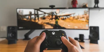 نمای اول شخص گیمینگ با کنترلر ایکس‌باکس روی میز، نمایشگر بزرگ با تصویر بازی اکشن، اسپیکرهای استودیویی و تزئینات شلف در پس‌زمینه، تمرکز بر تجربه بازی و تجهیزات گیمینگ