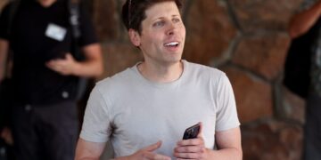 سم آلتمن (Sam Altman) در فضای باز در حالی که یک گوشی هوشمند در دست دارد و به روبرو نگاه می‌کند.