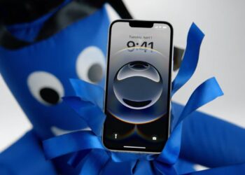 نمای جلوی یک آیفون با تاریخ اول آوریل که توسط یک عروسک بادی آبی رنگ نگه داشته شده است.