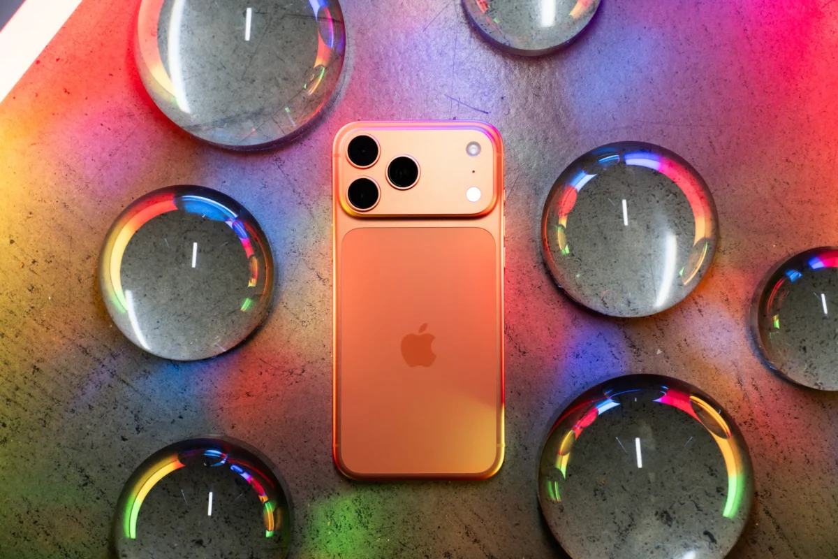 نمای پشت یک آیفون (احتمالاً مدل ۱۷ یا ایر) به رنگ مسی/نارنجی که روی سطحی باف‌دار و در میان گوی‌های شیشه‌ای شفاف قرار گرفته است.