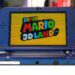 کنسول دستی Nintendo 3DS با بدنه آبی، در حالت باز، با نمایش صفحه عنوان بازی Super Mario 3D Land روی نمایشگر بالایی