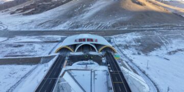 نمای هوایی از مجموعه ورودی و تأسیسات تونل تیان‌شان شِنگلی در منطقه کوهستانی سین‌کیانگ، با سازه‌های صنعتی، مسیرهای دسترسی و دامنه‌های پوشیده از برف.