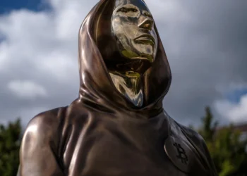 مجسمه برنزی با طراحی مفهومی از ساتوشی ناکاموتو در فضای باز، با لباس شنل‌مانند و نماد بیت‌کوین روی سینه، نماد هویت ناشناس خالق بیت‌کوین