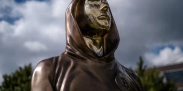 مجسمه برنزی با طراحی مفهومی از ساتوشی ناکاموتو در فضای باز، با لباس شنلمانند و نماد بیتکوین روی سینه، نماد هویت ناشناس خالق بیتکوین