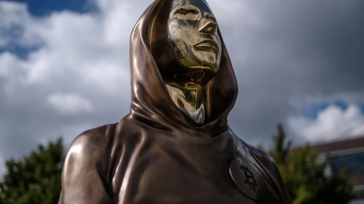 مجسمه برنزی با طراحی مفهومی از ساتوشی ناکاموتو در فضای باز، با لباس شنلمانند و نماد بیتکوین روی سینه، نماد هویت ناشناس خالق بیتکوین