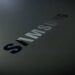 نمای نزدیک و زاویهدار از لوگوی نوشتاری سامسونگ با بافت تیره و براق روی یک سطح خاکستری.