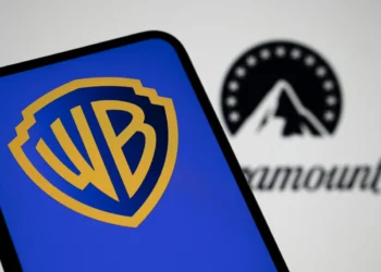 نمای نزدیک از صفحه‌نمایش گوشی هوشمند با نمایش لوگوی Warner Bros (WB) در رنگ آبی و زرد؛ در پس‌زمینه تار، لوگوی Paramount به‌صورت خارج از فوکوس قابل مشاهده است.