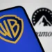 نمای نزدیک از صفحه‌نمایش گوشی هوشمند با نمایش لوگوی Warner Bros (WB) در رنگ آبی و زرد؛ در پس‌زمینه تار، لوگوی Paramount به‌صورت خارج از فوکوس قابل مشاهده است.