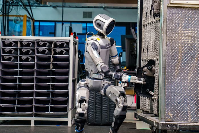 ربات‌های انسان‌نما در محیط کارخانه هیوندای، در حال کار با تجهیزات صنعتی و انجام وظیفه جابه‌جایی و تنظیم قطعات