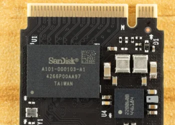 نمای بسیار نزدیک (ماکرو) از یک برد الکترونیکی حافظه فلش که تراشه اصلی SanDisk روی آن نصب شده است. مسیرهای مدار چاپی (PCB traces)، پین‌های اتصال طلایی و اجزای ریز الکترونیکی اطراف چیپ به‌وضوح دیده می‌شوند. تصویر روی یک سطح زرد بافت‌دار ثبت شده است.