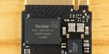 نمای بسیار نزدیک (ماکرو) از یک برد الکترونیکی حافظه فلش که تراشه اصلی SanDisk روی آن نصب شده است. مسیرهای مدار چاپی (PCB traces)، پین‌های اتصال طلایی و اجزای ریز الکترونیکی اطراف چیپ به‌وضوح دیده می‌شوند. تصویر روی یک سطح زرد بافت‌دار ثبت شده است.