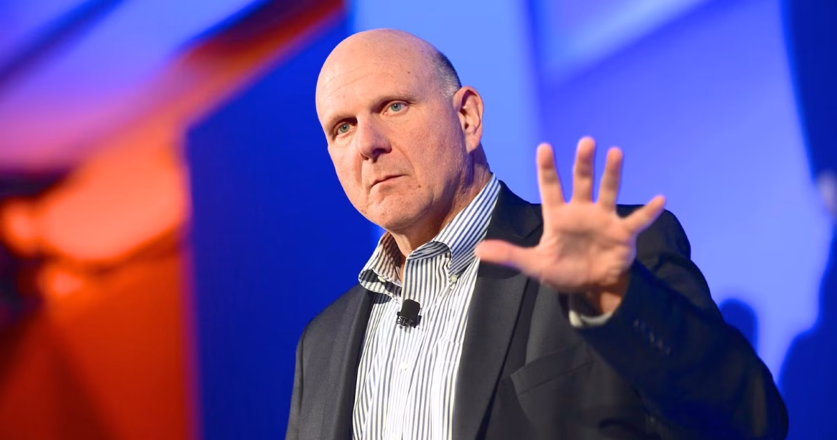 استیو بالمر در حال سخنرانی با کت و پیراهن راه راه، در حالی که دست خود را به نشانه عدد پنج بالا آورده است.