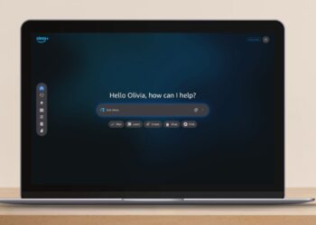 یک لپ‌تاپ باز روی میز چوبی که صفحه اصلی دستیار هوشمند جدید آمازون (Alexa.com) را نمایش می‌دهد.