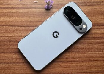 نمای پشت یک گوشی گوگل پیکسل (Google Pixel) با رنگ روشن که بر روی یک میز چوبی در کنار یک گل بنفش کوچک قرار دارد.