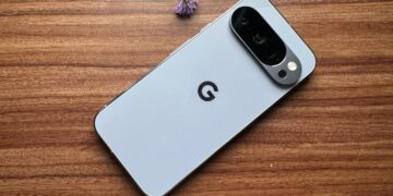 نمای پشت یک گوشی گوگل پیکسل (Google Pixel) با رنگ روشن که بر روی یک میز چوبی در کنار یک گل بنفش کوچک قرار دارد.
