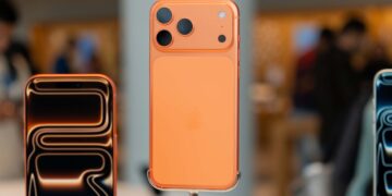 نمای نزدیک از گوشی Apple iPhone 17 Pro در رنگ نارنجی متالیک، نشان‌دهنده پنل پشتی با چینش سه‌گانه دوربین و فلاش LED در بخش بالا سمت چپ؛ نمایشگر فعال دستگاه در جلو با جلوه نوری انتزاعی و پشت‌زمینه تار فروشگاه.