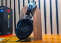 هدست ایسوس ROG Kithara قرار گرفته بر روی یک پایه چوبی عمودی در کنار جعبه محصول با پس‌زمینه دیواره آکوستیک.