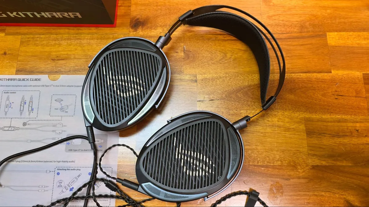 هدست گیمینگ ROG Kithara با کاپهای بیضیشکل و کابلهای بافته شده روی سطح چوبی در کنار دفترچه راهنما.