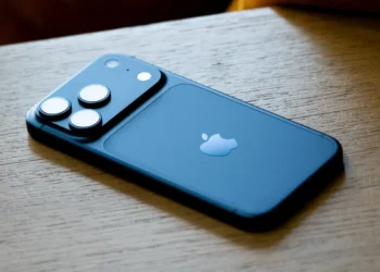 نمای پشتی یک گوشی iPhone با بدنه آبی مات که روی سطح چوبی قرار گرفته است؛ لوگوی Apple در مرکز پنل پشتی دیده میشود و ماژول دوربین مربعی شامل سه لنز برجسته و فلش LED با نورپردازی طبیعی و فوکوس کمعمق ثبت شده است.