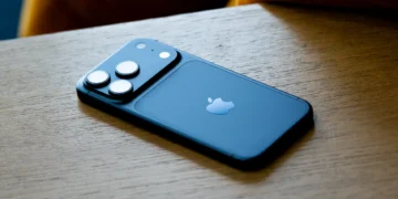 نمای پشتی یک گوشی iPhone با بدنه آبی مات که روی سطح چوبی قرار گرفته است؛ لوگوی Apple در مرکز پنل پشتی دیده می‌شود و ماژول دوربین مربعی شامل سه لنز برجسته و فلش LED با نورپردازی طبیعی و فوکوس کم‌عمق ثبت شده است.