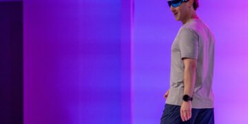 مارک زاکربرگ در حال قدم زدن روی صحنه با عینک هوشمند، تی‌شرت طوسی و شورت ورزشی تیره.