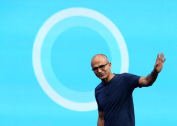 ساتیا نادلا، مدیرعامل مایکروسافت، در حال لبخند زدن و دست تکان دادن در مقابل یک پس‌زمینه آبی با لوگوی دایره‌ای سفید.