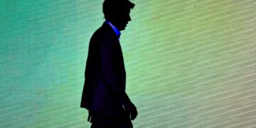 سایه تیره فردی (سام آلتمن) در حال حرکت از مقابل یک نمایشگر روشن بزرگ.