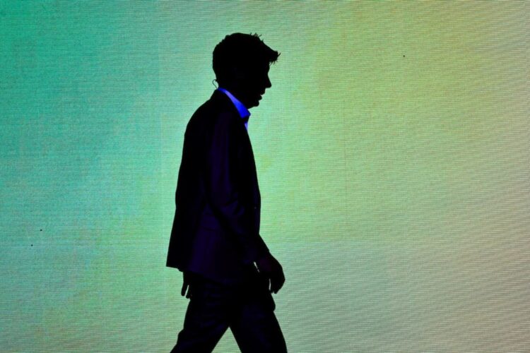 سایه تیره فردی (سام آلتمن) در حال حرکت از مقابل یک نمایشگر روشن بزرگ.