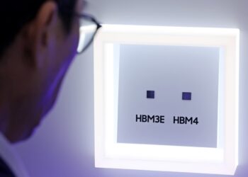 نمای نزدیک از فردی که در حال مشاهده دو تراشه کوچک حافظه با برچسب‌های HBM3E و HBM4 در یک محفظه نمایشگاهی روشن است.