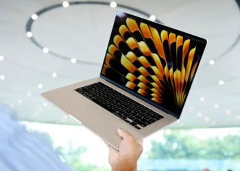 شخصی که یک لپ‌تاپ مک‌بوک ایر با والپیپر نارنجی را در یک فضای مدرن با سقف دایره‌ای نگه داشته است.