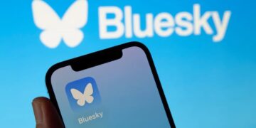 گوشی هوشمندی که آیکون اپلیکیشن Bluesky را نمایش می‌دهد و لوگوی بزرگ Bluesky در پس‌زمینه آن دیده می‌شود.