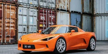 نمای جانبی خودروی الکتریکی JMEV SC01 با رنگ نارنجی، پارکشده در محوطه صنعتی با کانتینرها