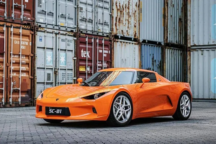 نمای جانبی خودروی الکتریکی JMEV SC01 با رنگ نارنجی، پارک‌شده در محوطه صنعتی با کانتینرها